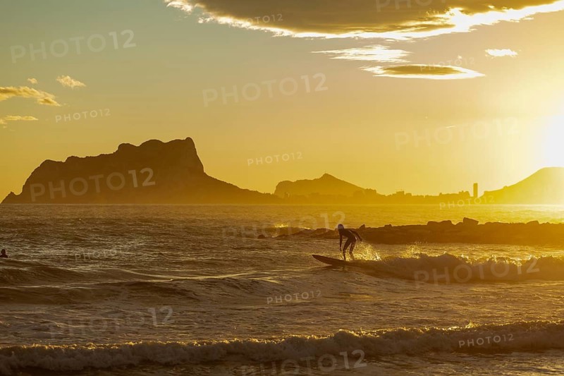 A surfer is silhouetted against the backdrop of a stunning sunset over the Mediterranean Sea off Costa Blanca. The peaceful water and vibrant sky create a breathtaking landscape