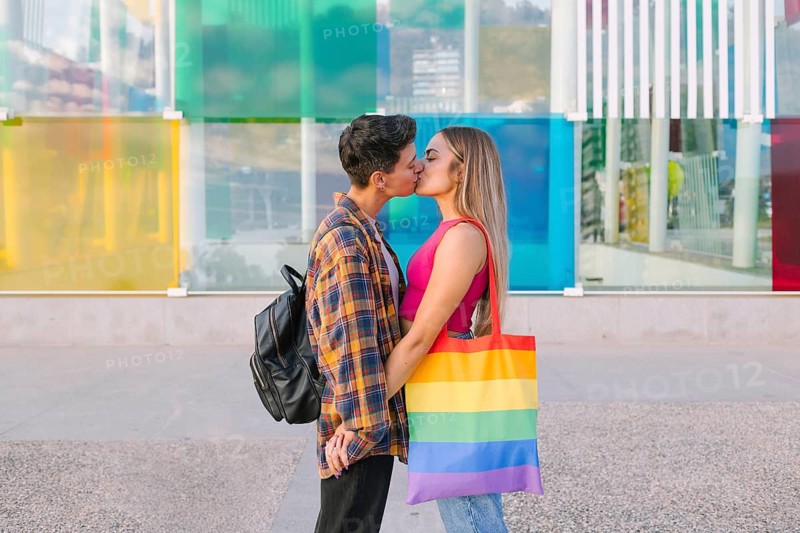 A joyful lesbian couple shares a tender kiss in front of a colorful city backdrop, holding a rainbow flag bag symbolizing pride. A celebration of love and diversity