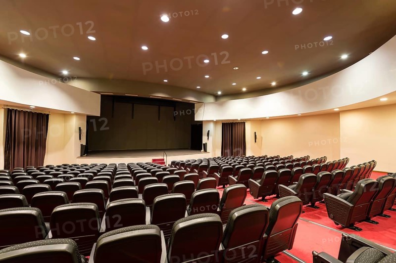 An empty convention center features rows of comfortable seats, bright stage lights, and elegant curtains. The spacious interior is designed for an immersive audience experience