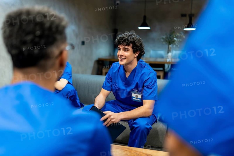 Young medical professional in blue scrubs listening and engaging in a discussion with team members during a brief break in a modern hospital or clinic environment