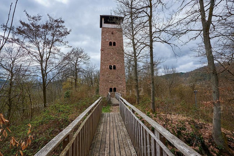 A stone observation tower stands at the end of a wooden bridge, surrounded by bare trees under a cloudy sky, Ohrsberg Tower, Eberbach, Odenwald, Rhein-Neckar district, Baden-Württemberg, Germany