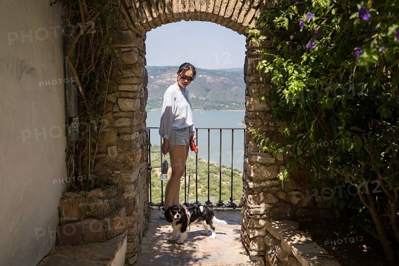 An Asian woman enjoys a sunny day with her dog in Castellar de la Frontera, Cadiz. Standing under a stone archway, she overlooks a beautiful landscape and serene water