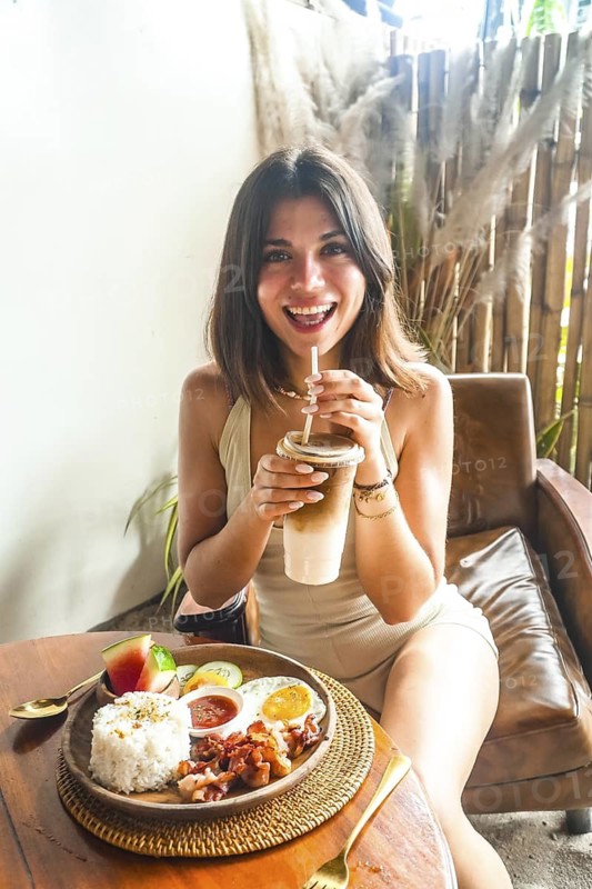 Happy tourist sipping iced coffee while enjoying a traditional filipino breakfast of bacon, eggs, rice, and fresh fruit in siargao