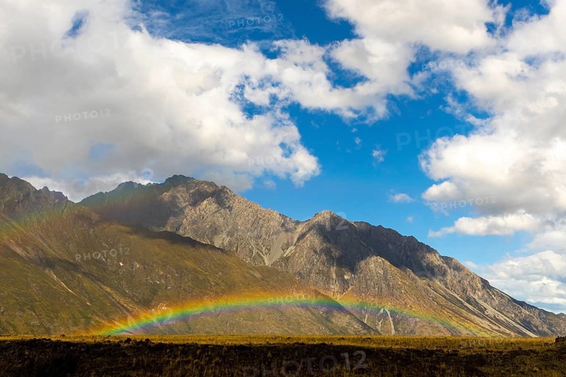 Scenic view of a vibrant rainbow stretching over Mount Cook in New Zealand during autumn. The mountain landscape is under a clear blue sky with scattered clouds, capturing nature beauty