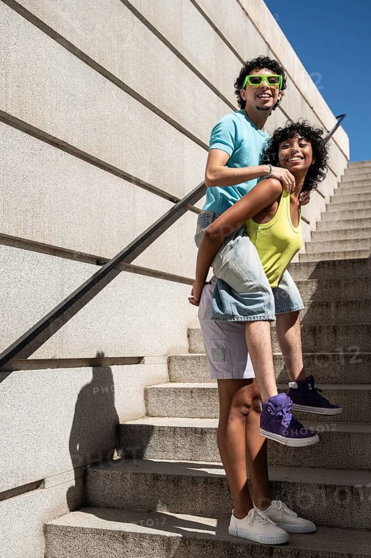A joyful multiethnic LGBTQ+ couple shares a lighthearted moment on outdoor stairs. One carries the other on their back, showcasing affection and happiness under a clear blue sky