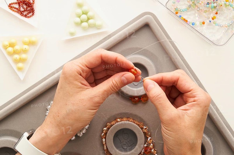 Top view of cropped unrecognizable woman's hands stringing beads on wire, creating handmade jewelry. Workspace filled with colorful materials, showcasing artistic creativity