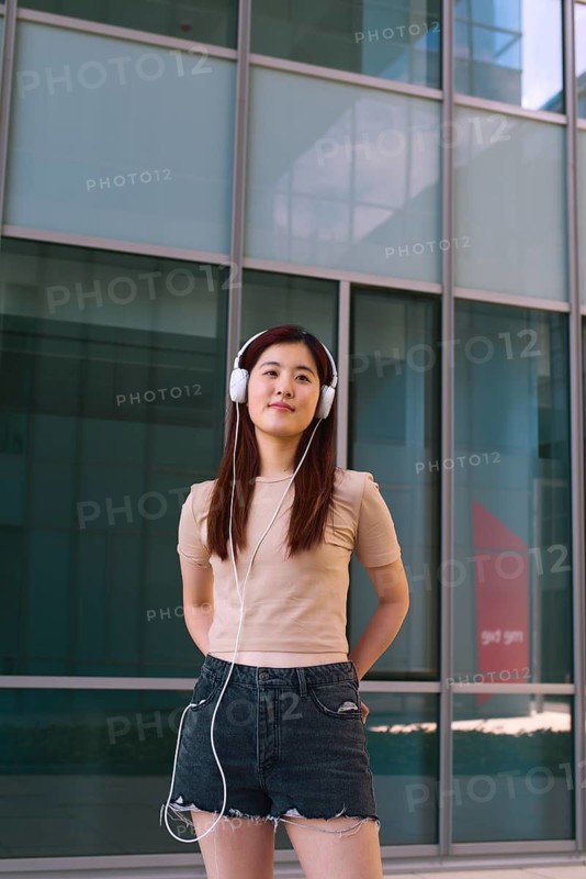 A young woman stands confidently outside a modern building, listening to music on white headphones. She exudes relaxation and enjoyment in her casual summer attire