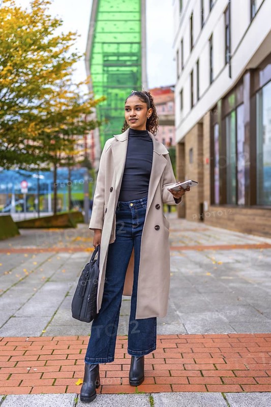 Vertical portrait of a stylish african businesswoman standing in the city holding smartphone and handbag