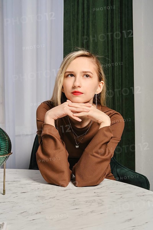 A young woman with blonde hair and red lipstick rests her chin on her hands while sitting at a marble table. Her fashion choice reflects elegance, complemented by a soft green backdrop