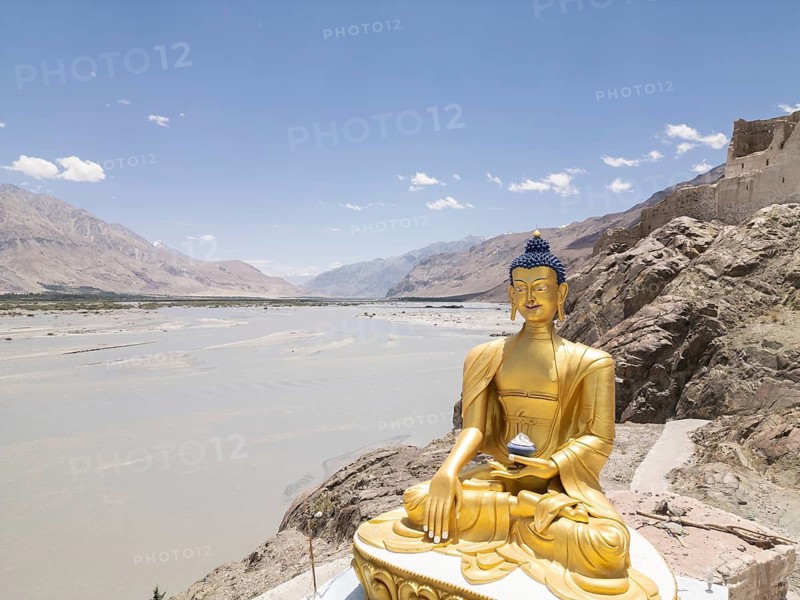 A large golden Buddha statue sits majestically on a rocky outcrop, overlooking the expansive Nubra Valley in Charasa, India, under a clear blue sky