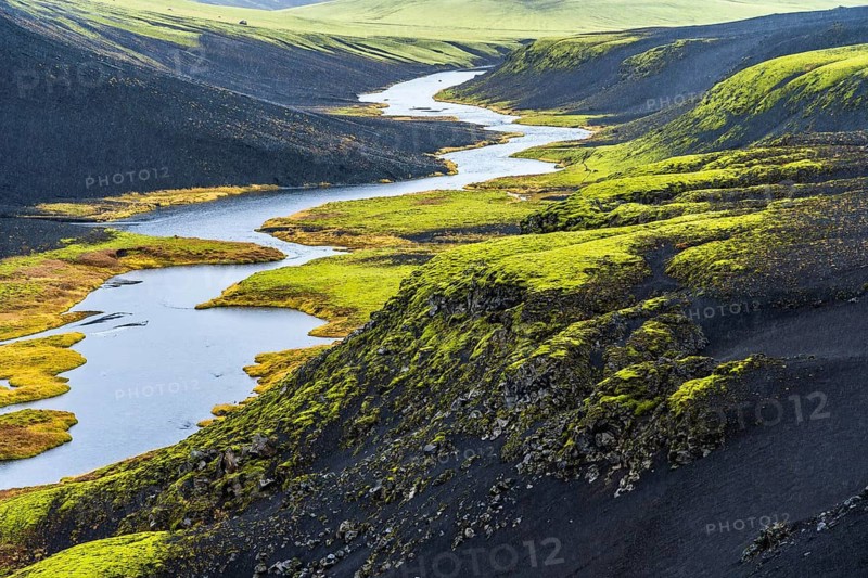 Aerial view of a winding river in Iceland, flanked by vibrant green moss-covered hills and rugged black rocks, showcasing the natural beauty and textures of this unique landscape