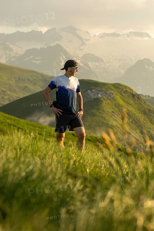 A man pauses while trail running on a scenic path surrounded by lush green hills and mountain views. He stands with hands on hips, taking in the serene natural environment