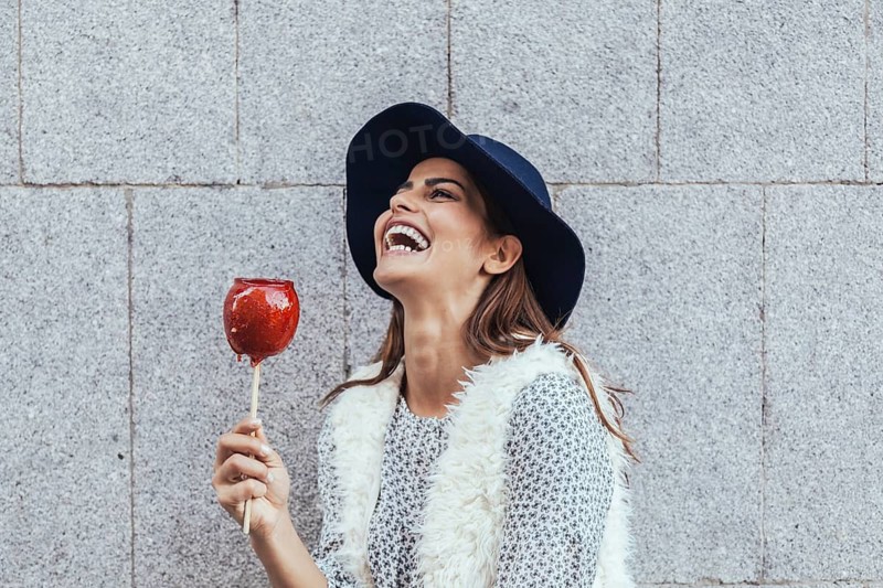A happy woman in a stylish hat and furry vest laughs joyfully while holding a candy apple. The grey stone backdrop adds contrast to her vibrant appearance