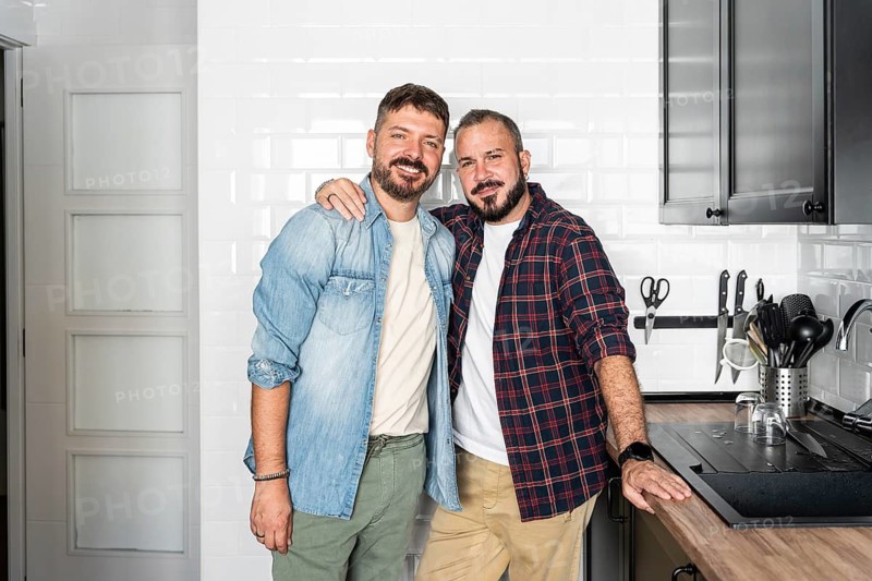 A joyful, loving gay couple stands in their modern kitchen, smiling warmly. Light shines through the room, highlighting their connection and stylish surroundings