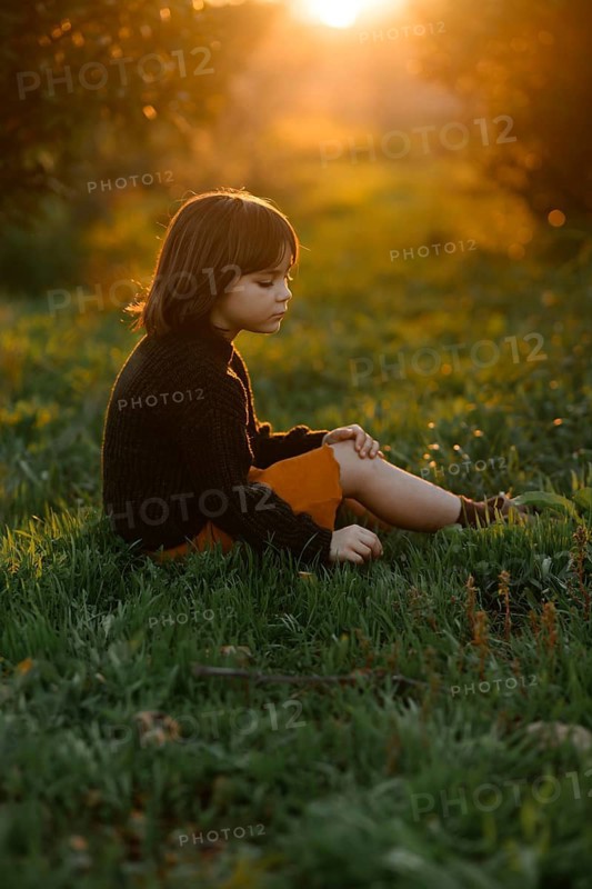 A child sits serenely in a grassy field with orange trees during sunset. The golden light creates a tranquil atmosphere, highlighting the child contemplative mood and the natural surroundings