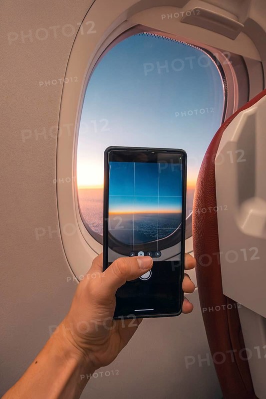 Cropped unrecognizable person captures a sunset over the ocean using a smartphone, viewed through an airplane window