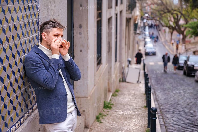 Man musician in a blue blazer and white sweater playing blues on a harmonica with eyes closed, standing outdoors in city street