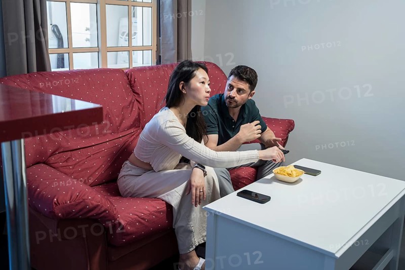 A diverse couple sits on a comfortable red sofa in their home, sharing a casual moment together while enjoying some snacks. They appear engaged in conversation, creating a warm, cozy atmosphere