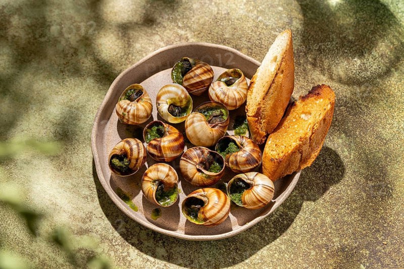Baked escargot snug in shells with lush green parsley and golden melted butter, served with slices of crusty toasted bread on an appealing ceramic plate