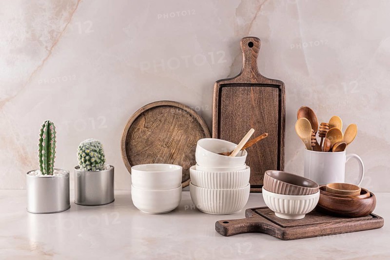 A well-arranged kitchen countertop featuring rustic wooden boards, ceramic bowls, wooden utensils, and two potted cacti against a marble backdrop