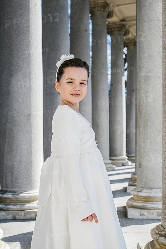 A young girl elegantly dressed in a First Communion Day dress stands in front of classic stone columns. The serene setting and attire reflect a moment of grace and tradition