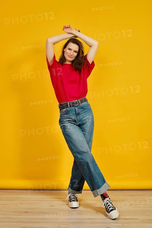 An adult woman stands confidently with her arms raised, wearing a red t-shirt and blue jeans