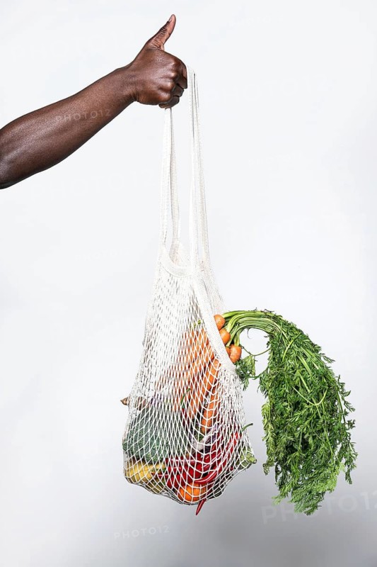 A person gives a thumbs-up while holding a mesh bag filled with fresh vegetables, symbolizing eco-friendly shopping and healthy living on a light background