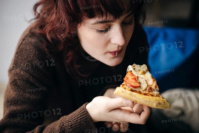 A young adult woman with curly red hair enjoys a delicious slice of pizza, focusing intently on her meal in a relaxed indoor environment