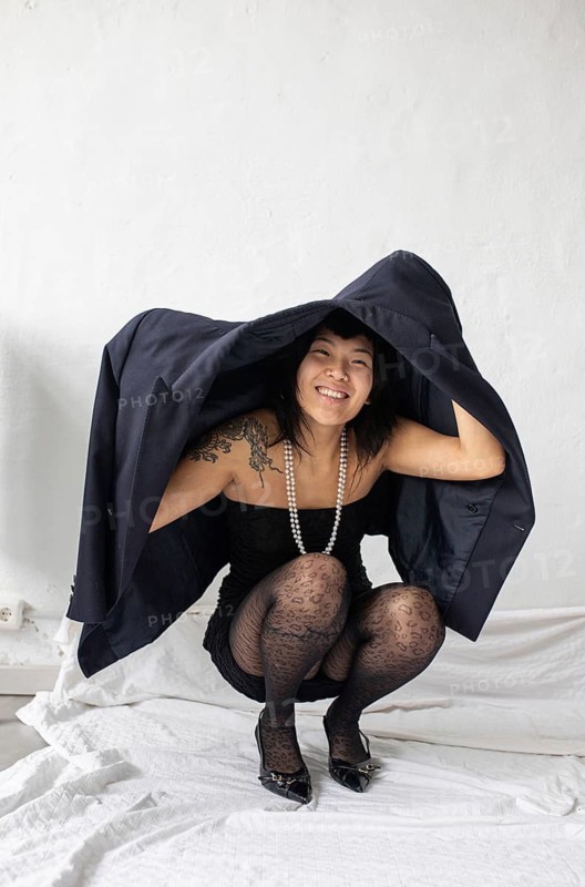 A cheerful Asian woman with meaningful tattoos crouches under a black jacket, laughing joyfully. She wears a stylish outfit with a pearl necklace, creating a playful portrait