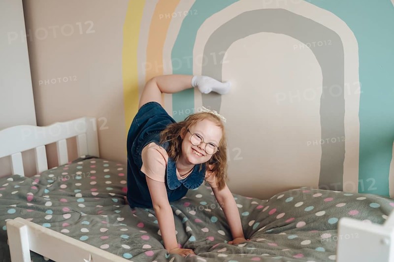 A joyful girl with Down syndrome plays on a bed in a colorful room with rainbow wall art. Her happy expression and dynamic pose capture a sense of fun and creativity
