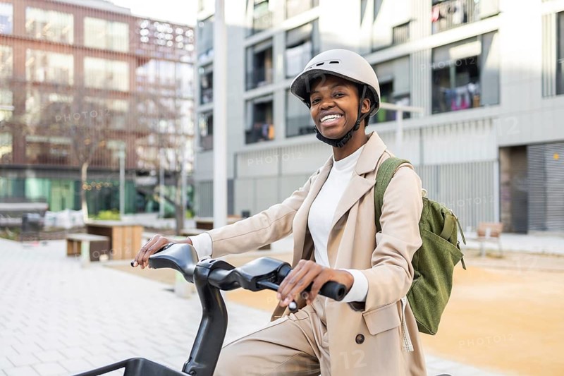 Black woman smiling while riding a bike in an urban city environment, wearing a helmet and backpack, showcasing modern and active lifestyle