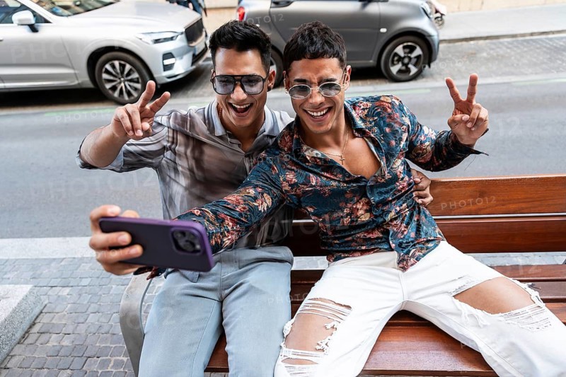 A cheerful latin gay couple takes a selfie on a sunny day in Madrid city, seated on a bench and flashing peace signs. They exude happiness and style in a vibrant urban setting