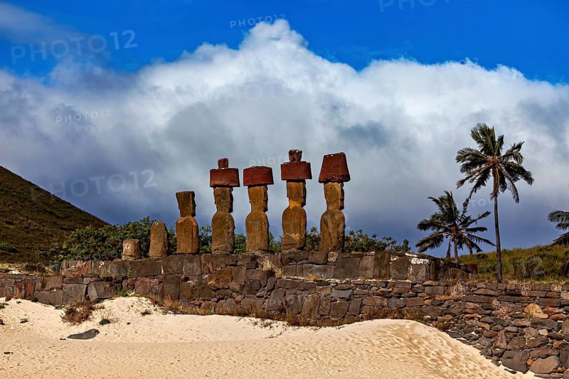 Stone statues on Easter Island against a cloudy blue sky surrounded by palm trees and sand, The Moai statues on Easter Island in the Pacific Ocean
