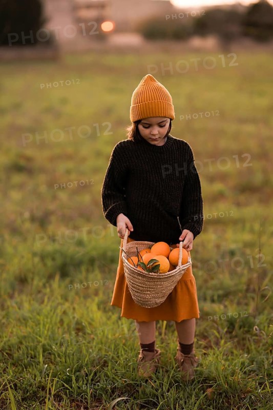 A child stands in a field, holding a basket filled with vibrant oranges. The scene conveys warmth and harvest, with the child dressed in cozy autumn attire