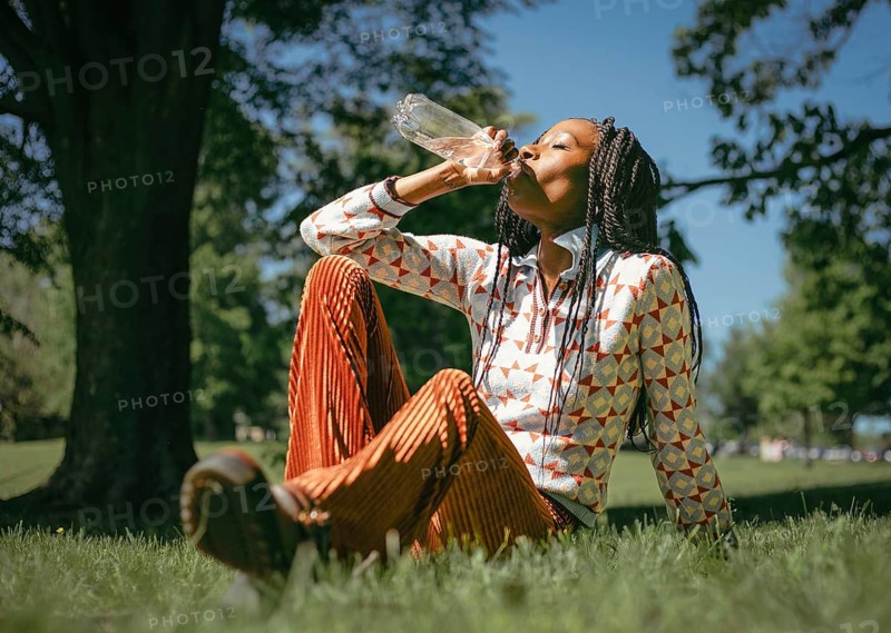 A woman sits on grass in a natural setting, savoring a refreshing drink of water. The sun shines through the trees, providing a serene and peaceful atmosphere ideal for relaxation