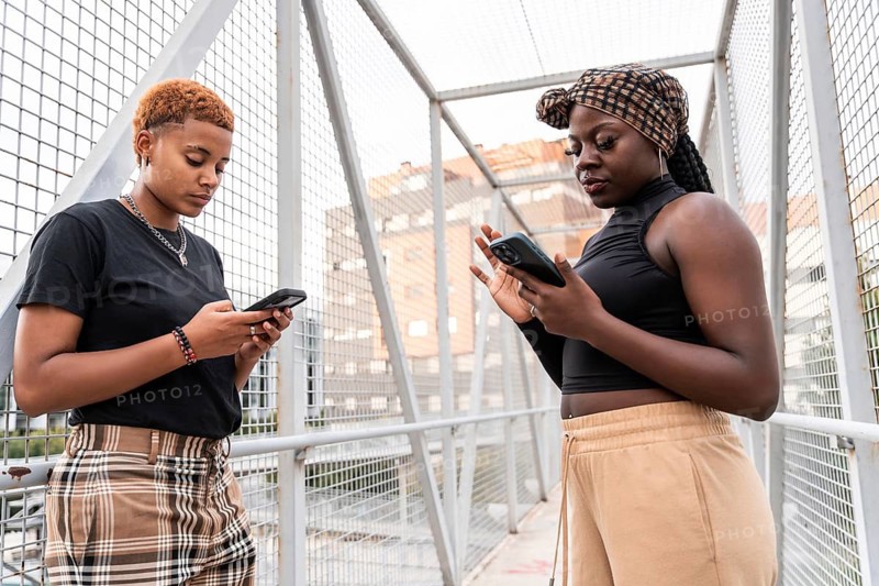 A multiethnic couple stands on a metal bridge, absorbed in their smartphones. They are casually dressed, highlighting modern urban life and connectivity