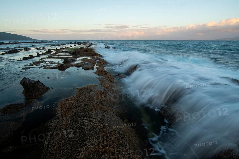 The sunset casts a warm glow over the rocky coastline of Tarifa, Cadiz. Waves crash against the shore, creating a dynamic and tranquil seascape scene