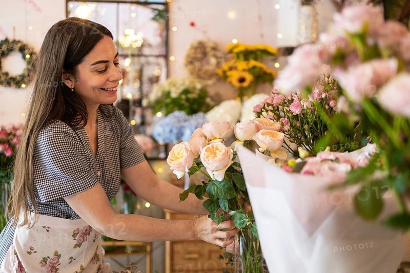 A woman with long hair and a checkered dress arranges delicate flowers in a picturesque flower shop. The shop is filled with colorful blooms and cozy, warm lighting