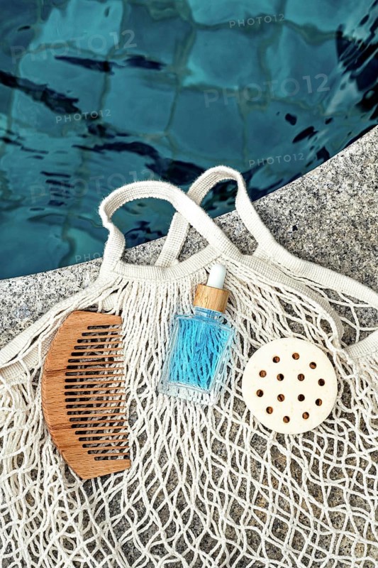 Top view image of a blue face care serum for men, set beside natural grooming tools, including a wooden comb and loofah, resting on a netted bag by the poolside, reflecting a serene ambiance