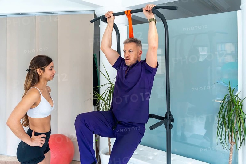 Male patient hanging from a bar doing leg raises for medical recovery and rehabilitation, with a female physiotherapist guiding the exercise for improved strength and range of motion