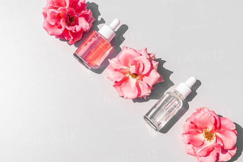 Top view of rose water and serum bottles accompanied by vibrant pink roses on a minimalist white surface, creating a fresh and elegant aesthetic