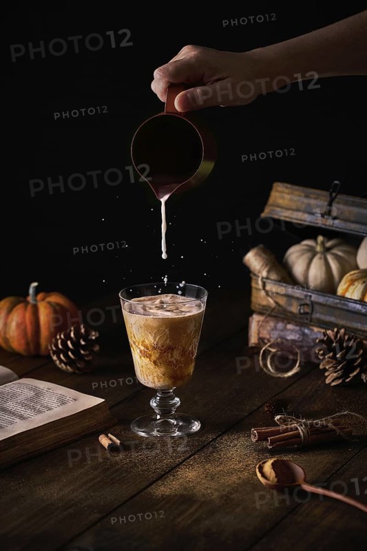 Crop anonymous person adding milk from small jug to glass with pumpkin spice latte at autumn decorative table