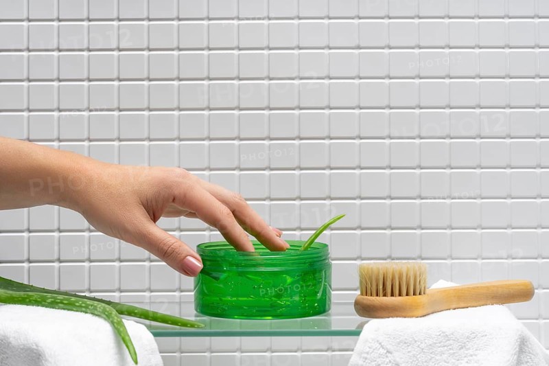 A female hand is shown scooping aloe gel from a jar in a tiled bathroom, alongside a body cream and wooden brush on a glass shelf