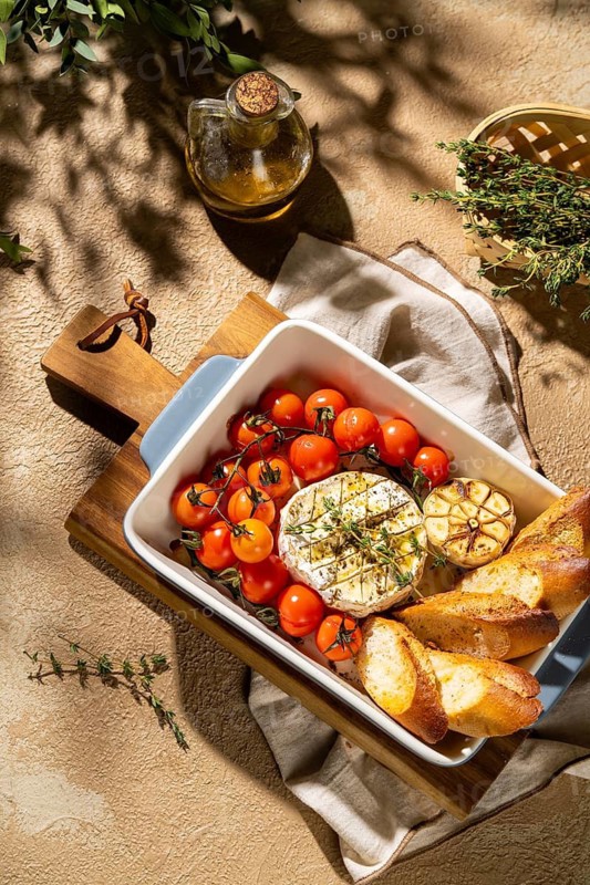 A rustic setting featuring baked Camembert cheese in a dish with cherry tomatoes, garlic, herbs, and crusty bread slices on a wooden board. Ideal for gourmet cuisine themes