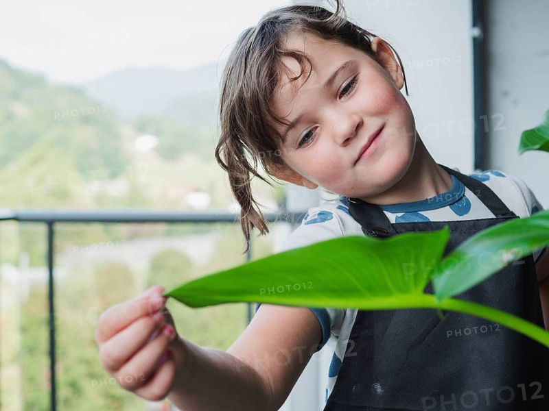 A young girl examines a large, healthy leaf on a balcony, with mountains visible in the background