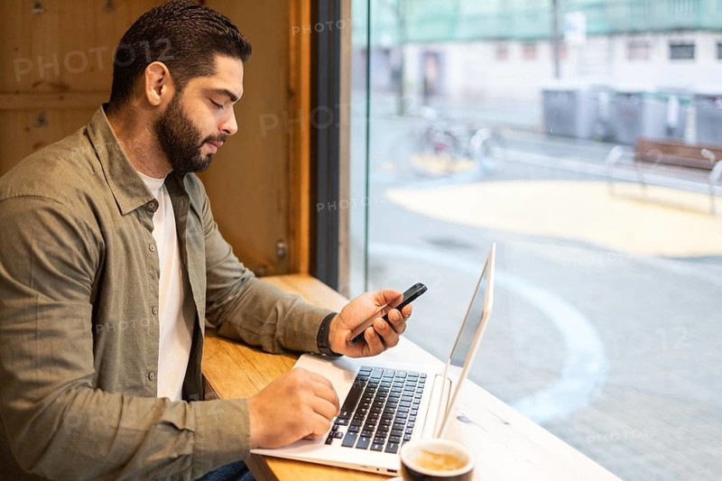 Focused individual working on a laptop while checking a smartphone in a cozy cafe setting of the city. A cup of coffee sits nearby, creating a relaxed yet productive atmosphere