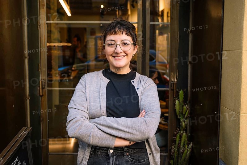 A friendly barista stands confidently at the entrance of a specialty coffee shop, exuding warmth and welcoming vibes. The inviting atmosphere is perfect for coffee enthusiasts