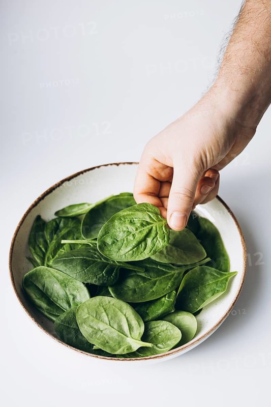 A hand reaches into a bowl of fresh green spinach leaves, set against a clean white background. The vibrant greens evoke health, freshness, and natural goodness