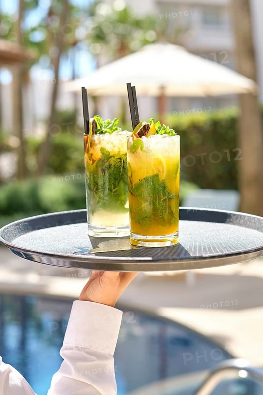 Two colorful cocktails with mint and lime on a tray, held by a waiter by the pool. The sunny backdrop features greenery and a parasol, epitomizing relaxation