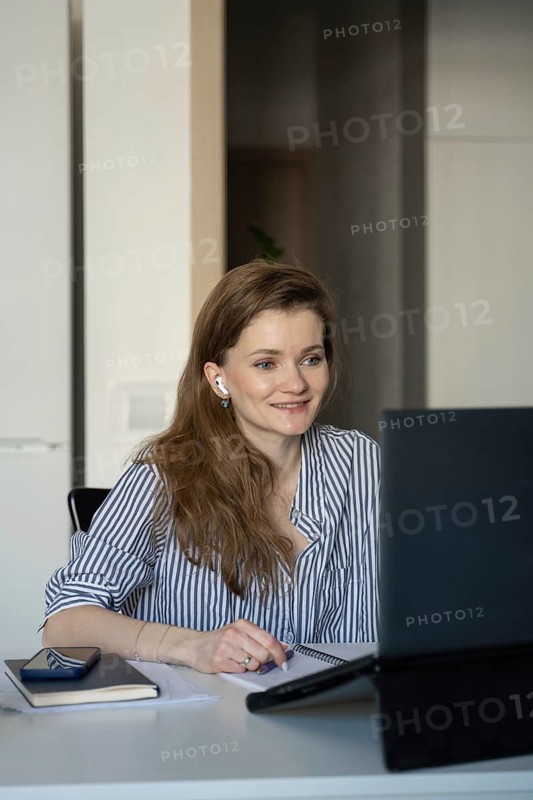 A woman sitting at her desk at home, smiling as she looks at her laptop screen. She is wearing earbuds, suggesting remote communication or entertainment